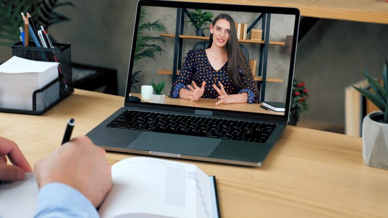 Prüfling auf Laptopbildschirm. Prüfende Person sitzt am Schreibtisch und macht sich Notizen.