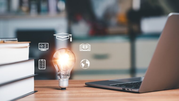 Schreibtisch mit Laptop und Büchern. Im Vordergrund eine Glühbirne, die Icons für digitale Bildung umgibt.