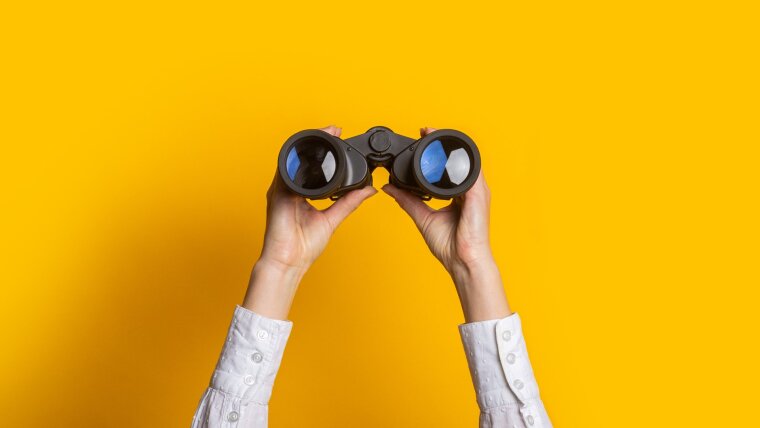 Zwei Hände halten ein Fernglas vor gelbem Hintergrund.